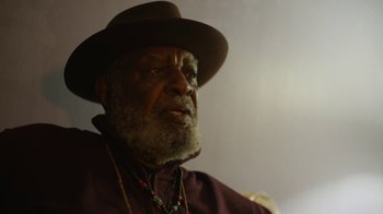 Movie still from “Full Circle” (2023), directed by Steven Soderbergh – An older man wearing a hat and a necklace; Close Up shot, Low angle