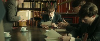 Movie still from “The Theory of Everything” (2014), directed by James Marsh – A man sitting at a table in front of two other people; Medium shot, Over the shoulder angle