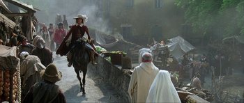 Movie still from “The Three Musketeers” (1993), directed by Stephen Herek – A woman riding a horse down a street with a man walking behind her; Wide shot, Over the shoulder angle