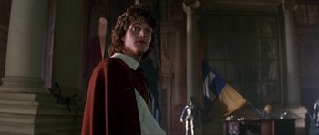 Movie still from “The Three Musketeers” (1993), directed by Stephen Herek – A man in a red coat standing next to a pillar; Medium shot, Over the shoulder angle