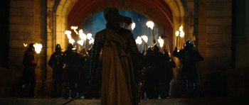 Movie still from “The Three Musketeers” (2011), directed by Paul W.S. Anderson – A person in a hooded cloak walking in front of a group of people holding torches; Wide shot, Over the shoulder angle