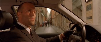 Movie still from “The Transporter” (2002), directed by Corey Yuen – A man in a suit driving a car in the street; Close Up shot, Low angle