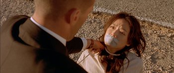 Movie still from “The Transporter” (2002), directed by Corey Yuen – A woman tied to the side of a road with a man holding her mouth; Close Up shot, Over the shoulder angle