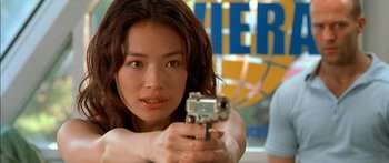 Movie still from “The Transporter” (2002), directed by Corey Yuen – A beautiful young woman holding a gun while standing in front of a basketball court; Close Up shot, Low angle