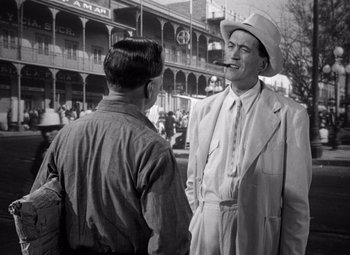 Movie still from “The Treasure of the Sierra Madre” (1948), directed by John Huston – A man in a suit and hat talking to another man in front of a building; Medium shot, Over the shoulder angle