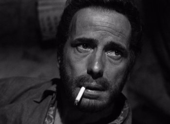 Movie still from “The Treasure of the Sierra Madre” (1948), directed by John Huston – A black and white photo of a man smoking a cigarette; Close Up shot, High angle