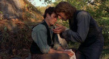 Movie still from “The Village” (2004), directed by M. Night Shyamalan – Two men are sitting in the woods and one of the men is holding his hand out to the other man; Medium shot, Over the shoulder angle