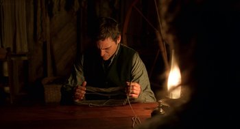 Movie still from “The Village” (2004), directed by M. Night Shyamalan – A man sitting at a wooden table knitting; Medium shot, Over the shoulder angle
