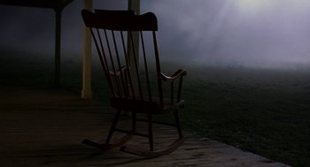 Movie still from “The Village” (2004), directed by M. Night Shyamalan – An empty rocking chair sitting on a porch in the fog; Wide shot, High angle