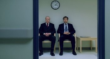 Movie still from “The Weather Man” (2005), directed by Gore Verbinski – Two businessmen sitting in chairs in front of a wall clock; Wide shot, Low angle