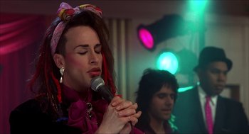 Movie still from “The Wedding Singer” (1998), directed by Frank Coraci – A man with a pink bow on his head is holding a microphone; Close Up shot, Over the shoulder angle
