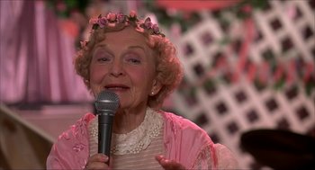 Movie still from “The Wedding Singer” (1998), directed by Frank Coraci – An older woman wearing a pink dress holding a microphone; Close Up shot, Over the shoulder angle