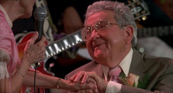 Movie still from “The Wedding Singer” (1998), directed by Frank Coraci – An older man wearing a suit and tie holding hands with a microphone; Close Up shot, Over the shoulder angle