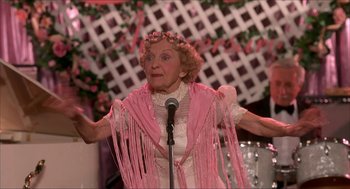 Movie still from “The Wedding Singer” (1998), directed by Frank Coraci – An older woman wearing a pink shawl and holding a microphone in front of her mouth; Medium shot, Low angle
