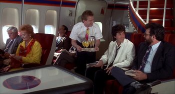 Movie still from “The Wedding Singer” (1998), directed by Frank Coraci – A group of people sitting on a plane with a tray of food on their lap; Medium shot, Over the shoulder angle