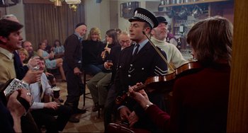 Movie still from “The Wicker Man” (1973), directed by Robin Hardy – A group of people sitting in a room with a man playing a guitar; Wide shot, High angle