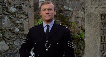 Movie still from “The Wicker Man” (1973), directed by Robin Hardy – A man in a uniform standing in front of a stone wall; Close Up shot, Low angle