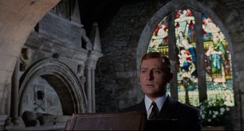 Movie still from “The Wicker Man” (1973), directed by Robin Hardy – A man in a suit and tie in front of a stained glass window; Medium shot, Low angle