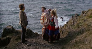 Movie still from “The Wicker Man” (1973), directed by Robin Hardy – A man and two women dressed in costumes near the ocean; Wide shot, High angle