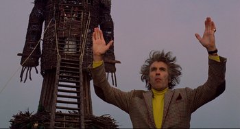 Movie still from “The Wicker Man” (1973), directed by Robin Hardy – A man in a yellow shirt and a plaid jacket waves; Medium shot, Low angle