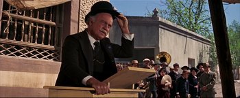 An older man in a suit and hat is holding his hat up