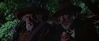 Movie still from “The Wild Bunch” (1969), directed by Sam Peckinpah – Two old men with a beard and a cowboy hat are sitting next to each other holding a jar; Close Up shot, Low angle