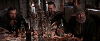 Movie still from “The Wild Bunch” (1969), directed by Sam Peckinpah – A man sitting at a dinner table with wine bottles and candles; Medium shot, Over the shoulder angle