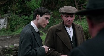 Movie still from “The Wind that Shakes the Barley” (2006), directed by Ken Loach – Two men standing next to each other while one smokes a cigarette; Medium shot, Over the shoulder angle