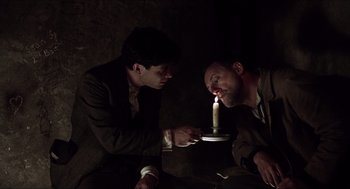 Movie still from “The Wind that Shakes the Barley” (2006), directed by Ken Loach – Two men are lighting a candle in the dark; Medium shot, Over the shoulder angle