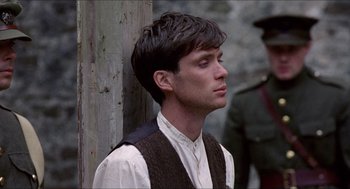Movie still from “The Wind that Shakes the Barley” (2006), directed by Ken Loach – A young man wearing a vest and standing next to a wooden post; Close Up shot, Low angle