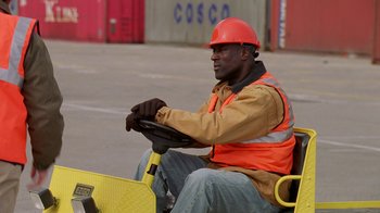 Movie still from “The Wire” (2002), created by David Simon – A man in an orange hard hat driving a yellow cart; Medium shot, High angle