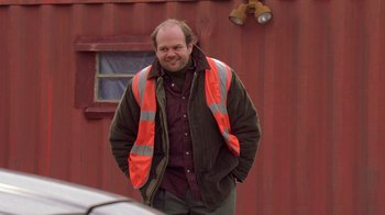 Movie still from “The Wire” (2002), created by David Simon – A man in a jacket and vest standing in front of a red building; Medium shot, Over the shoulder angle