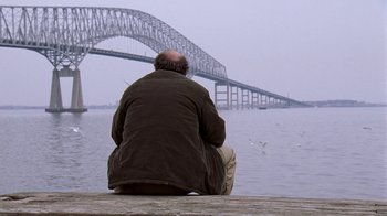 Movie still from “The Wire” (2002), created by David Simon – A man sitting on a pier looking at a bridge; Wide shot, Over the shoulder angle