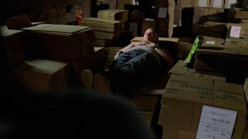 Movie still from “The Wire” (2002), created by David Simon – A man laying on the ground in a room filled with boxes; Wide shot, High angle