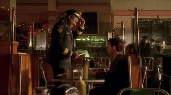 Movie still from “The Wire” (2002), created by David Simon – A man in a uniform is standing at a table with another man in a suit; Medium shot, Over the shoulder angle