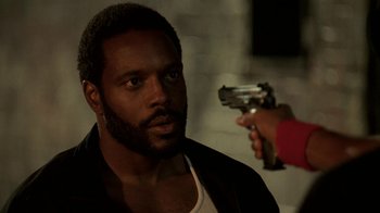 Movie still from “The Wire” (2002), created by David Simon – A man with a gun pointed at him in a dark room; Close Up shot, Over the shoulder angle