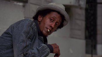 Movie still from “The Wire” (2002), created by David Simon – A man with a hat smoking a cigarette outside; Close Up shot, Low angle