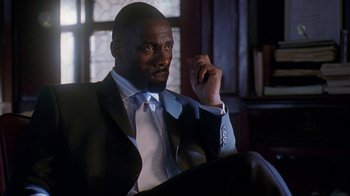 Movie still from “The Wire” (2002), created by David Simon – A man in a suit and tie sitting in front of a window; Medium shot, Low angle