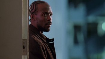 Movie still from “The Wire” (2002), created by David Simon – A black man with a beard and cornrows is standing in front of a building; Close Up shot, Low angle