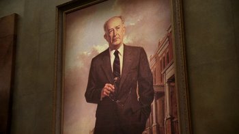 Movie still from “The Wire” (2002), created by David Simon – A painting of an older man in a suit and tie; Medium shot, Low angle