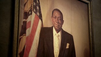 Movie still from “The Wire” (2002), created by David Simon – An older man wearing a suit and tie standing in front of an american flag; Extreme Close Up shot, Low angle