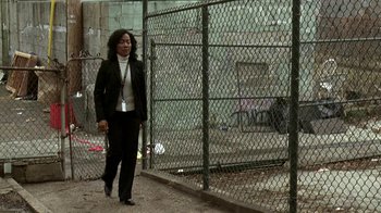 Movie still from “The Wire” (2002), created by David Simon – A woman in a black suit walking by a chain link fence; Wide shot, High angle