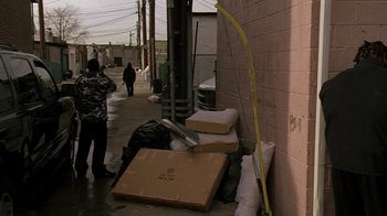 Movie still from “The Wire” (2002), created by David Simon – A person standing next to a wall with a box; Wide shot, Over the shoulder angle