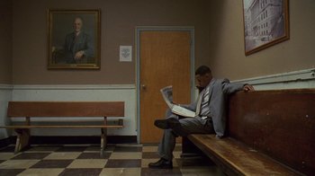 Movie still from “The Wire” (2002), created by David Simon – A man sitting on top of a wooden bench reading a newspaper; Wide shot, Low angle