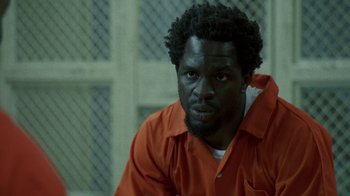 Movie still from “The Wire” (2002), created by David Simon – A man in an orange shirt sitting in front of a fence; Close Up shot, Over the shoulder angle