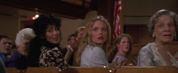 Movie still from “The Witches of Eastwick” (1987), directed by George Miller – A group of people sitting in front of a wooden table; Medium shot, Over the shoulder angle