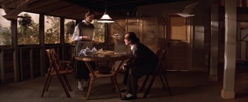 Movie still from “The Witches of Eastwick” (1987), directed by George Miller – A man sitting at a table with a woman standing next to him; Wide shot, High angle