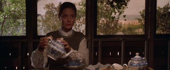 Movie still from “The Witches of Eastwick” (1987), directed by George Miller – A woman sitting at a table pouring tea from a tea pot into a tea cup; Medium shot, High angle