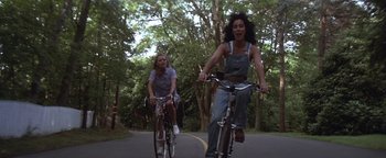 Movie still from “The Witches of Eastwick” (1987), directed by George Miller – Two women are riding bikes down the street; Wide shot, Low angle