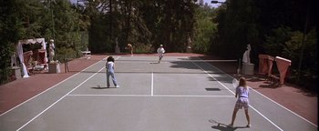 Movie still from “The Witches of Eastwick” (1987), directed by George Miller – A group of people on a tennis court playing a game; Extreme Wide shot, High angle
